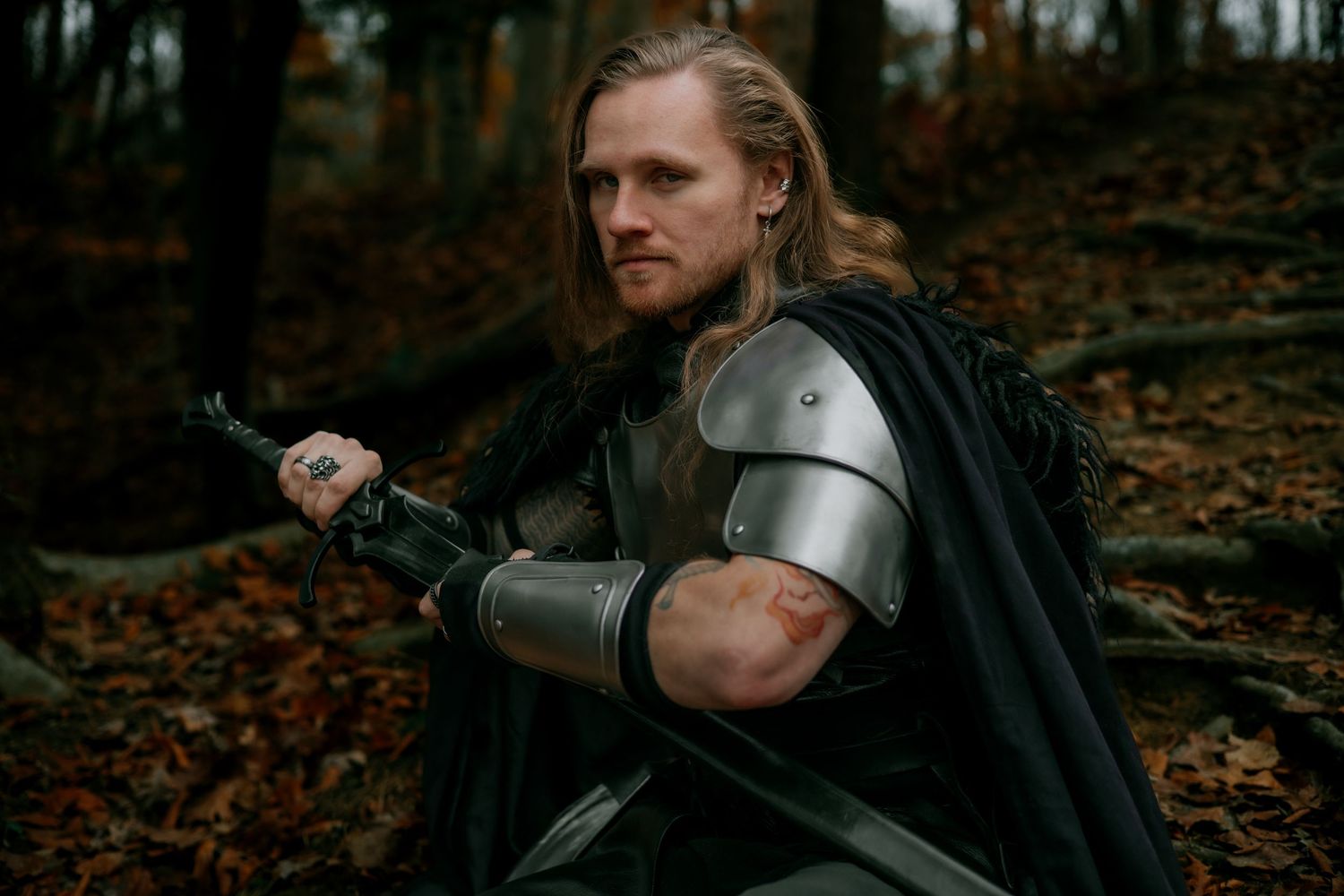 Medieval warrior in armor poses with sword among autumn leaves in atmospheric forest setting.
