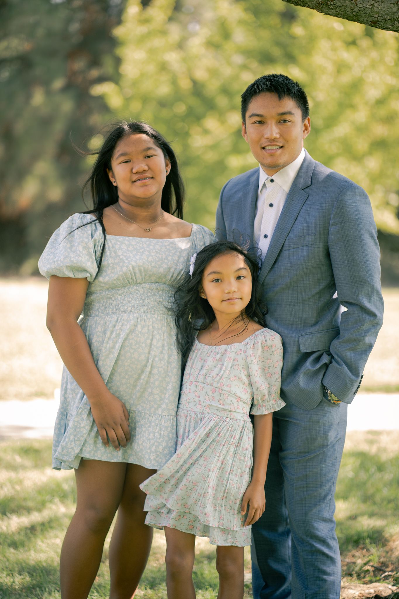 Family wearing coordinated outfits poses together outdoors in a natural setting with soft lighting and greenery.