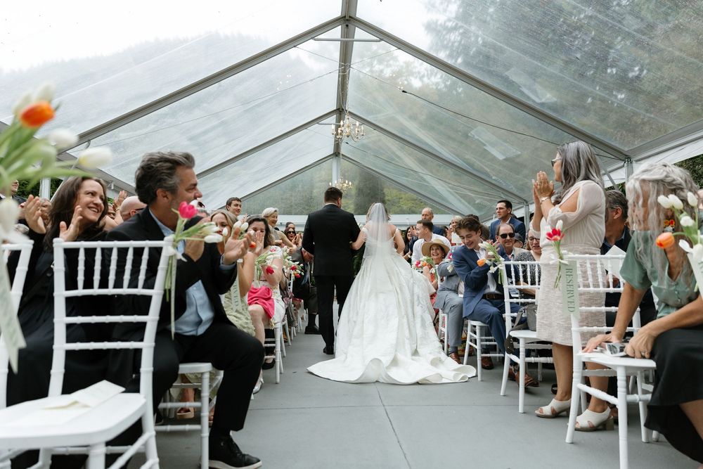 Wedding reception setup in a clear tent with white chairs and guests celebrating.