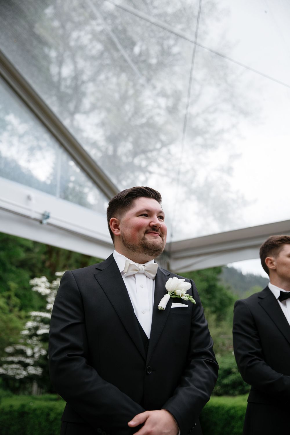 Groomsmen in black tuxedos and white bowties share a candid moment outdoors during a wedding celebration.