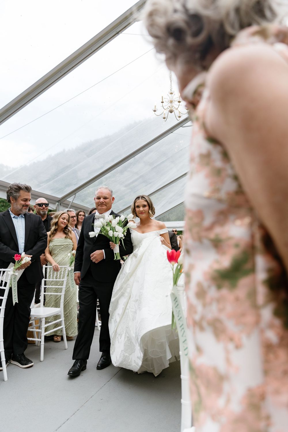 A wedding ceremony processional takes place under an elegant clear tent with white chairs and floral decorations.