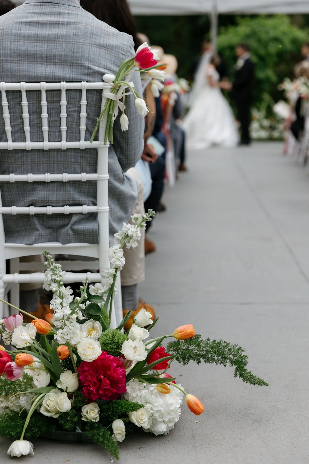 White chiavari chairs line an aisle decorated with colorful floral arrangements featuring red and orange blooms.