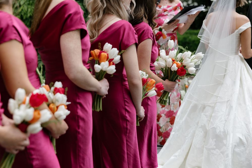 Bridesmaids in magenta dresses stand in line holding colorful floral bouquets at an outdoor wedding.