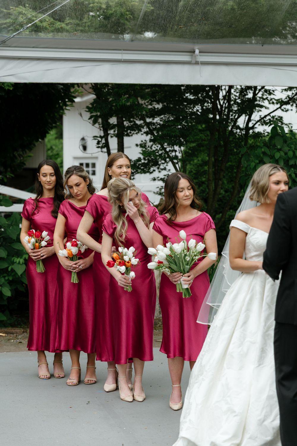 A wedding party in burgundy bridesmaid dresses and a white bridal gown pose together outdoors.