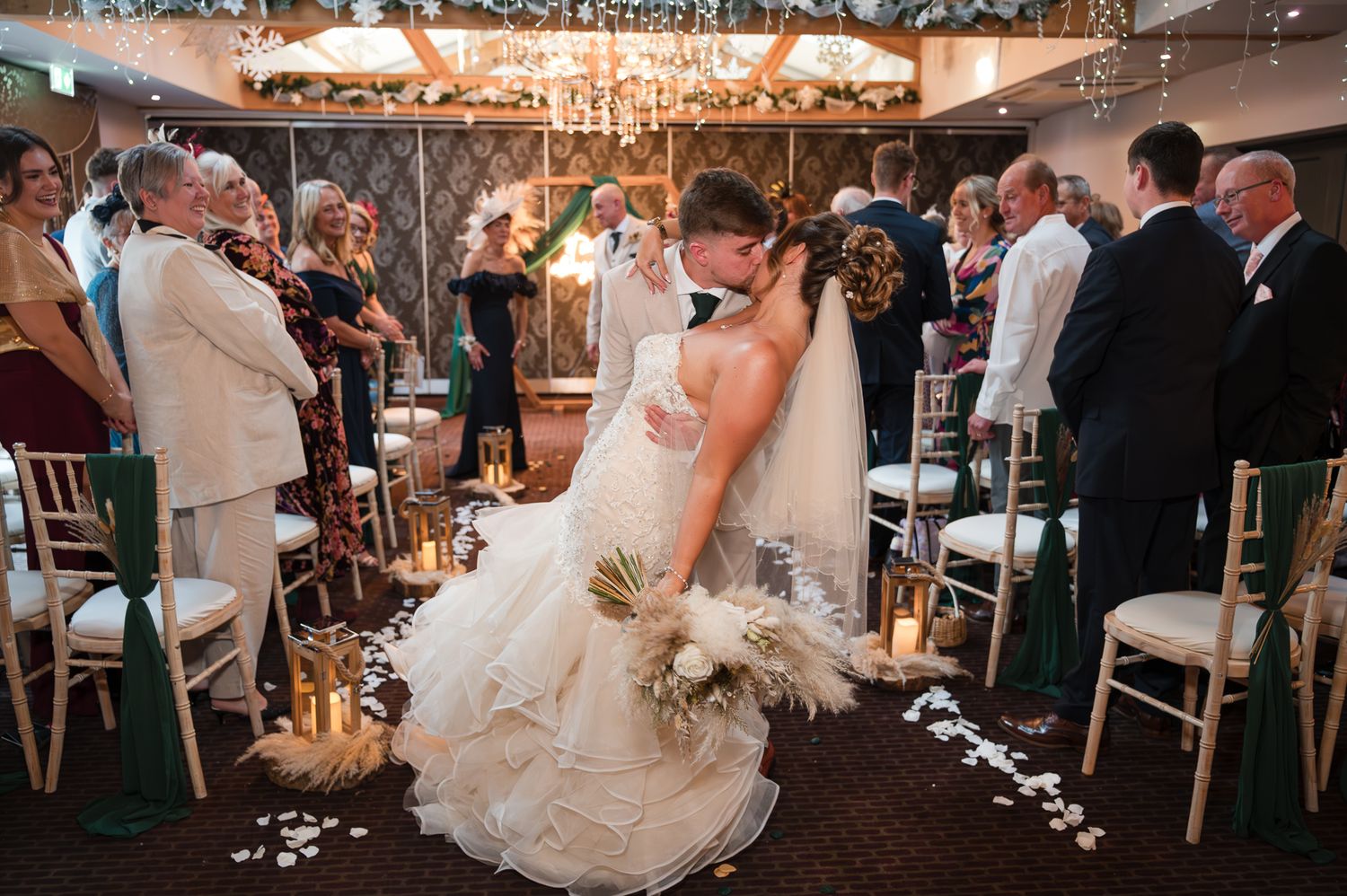 Wedding reception guests dance and celebrate under warm string lights in a rustic barn venue.