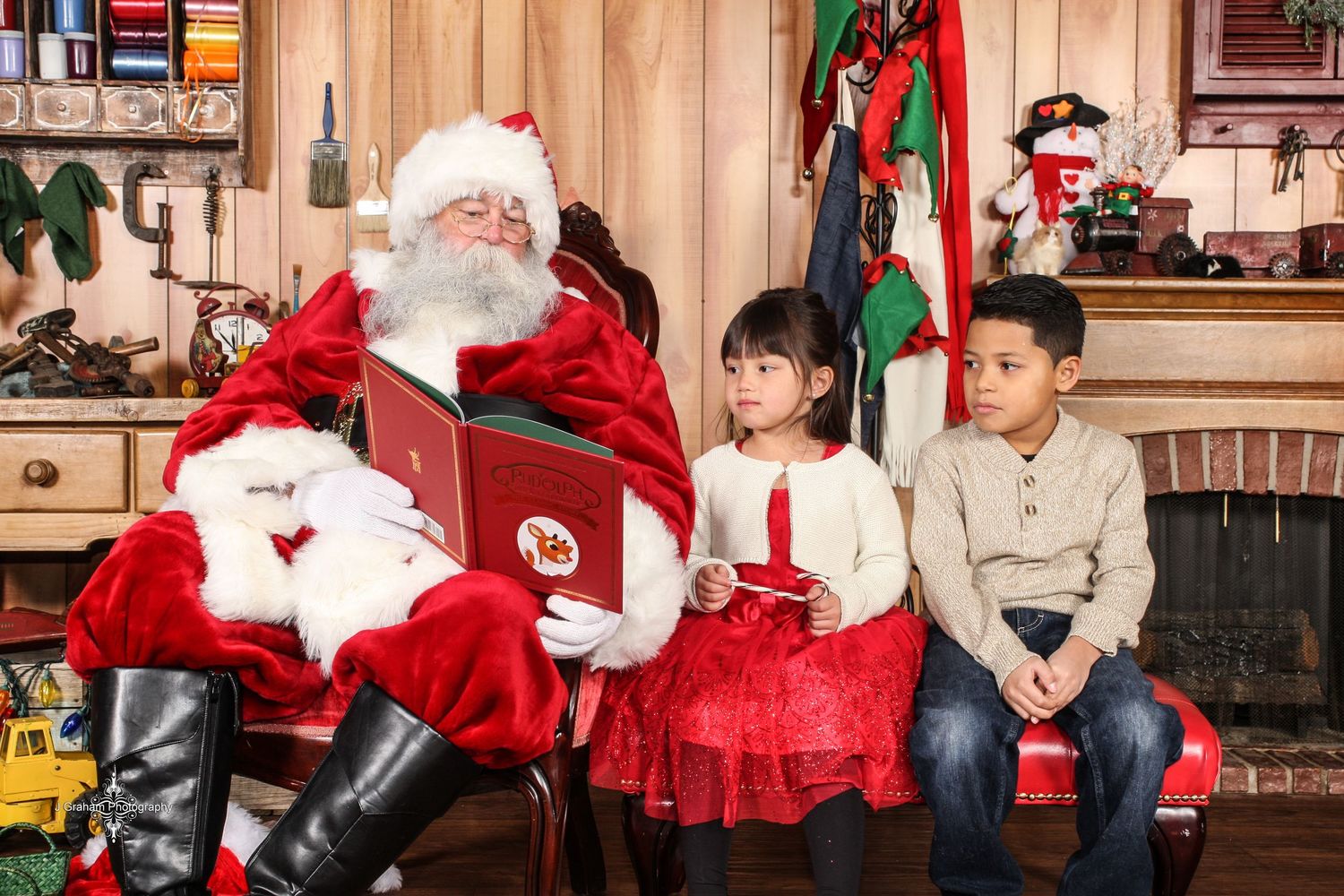 Two children in holiday pajamas open Christmas presents near a decorated tree while Santa sits on a red leather couch.