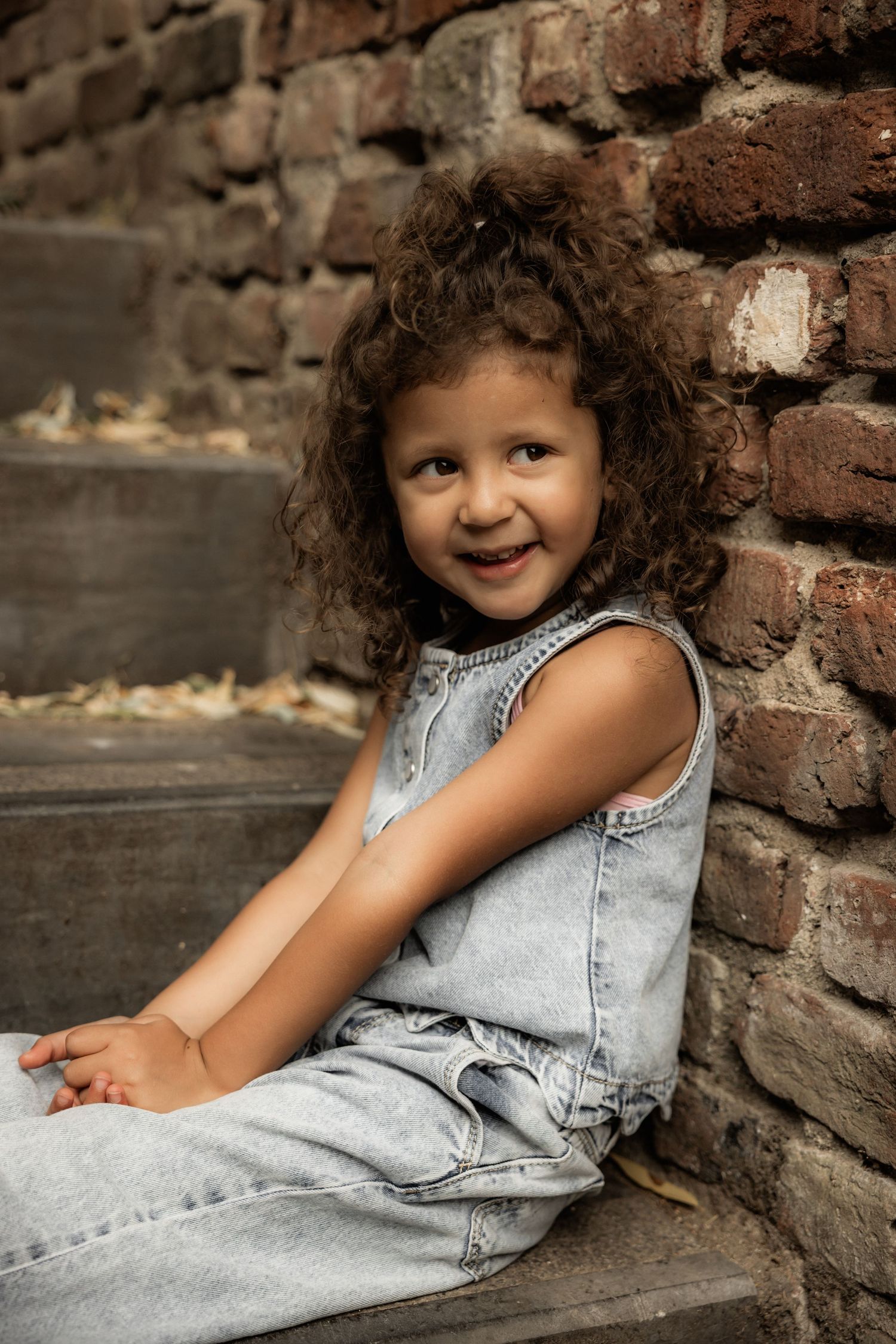 Child in denim clothing sitting against rustic brick wall with natural lighting.