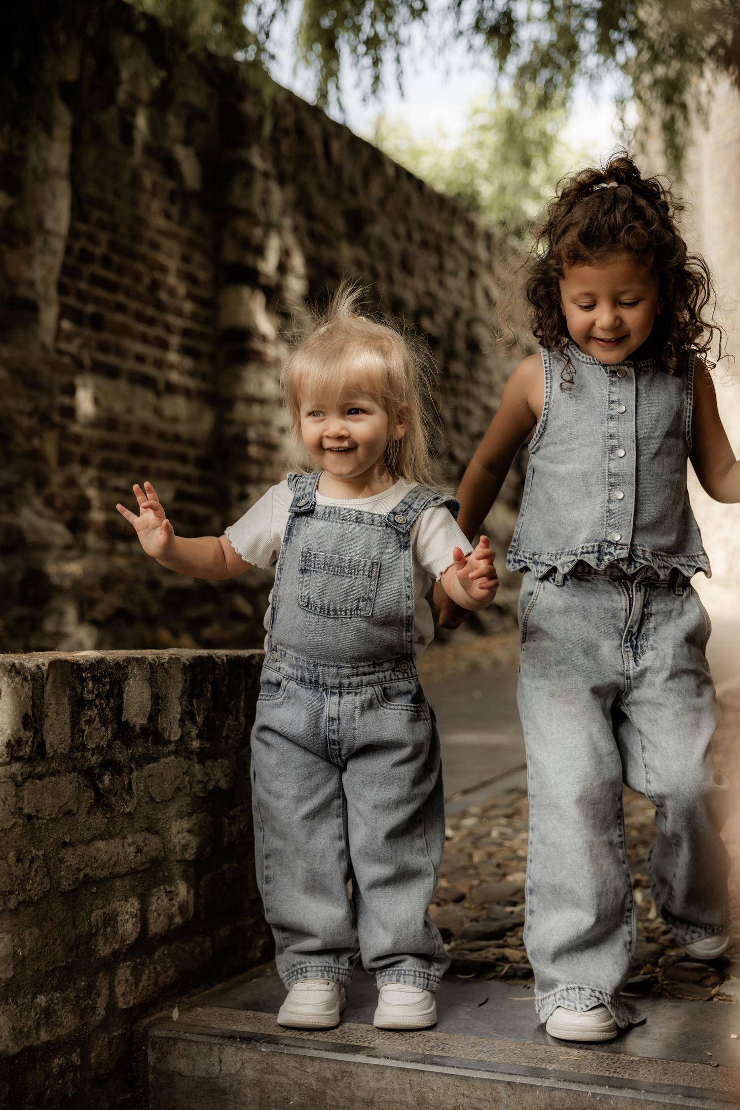 Two small children in matching denim overalls playing together against a brick wall.