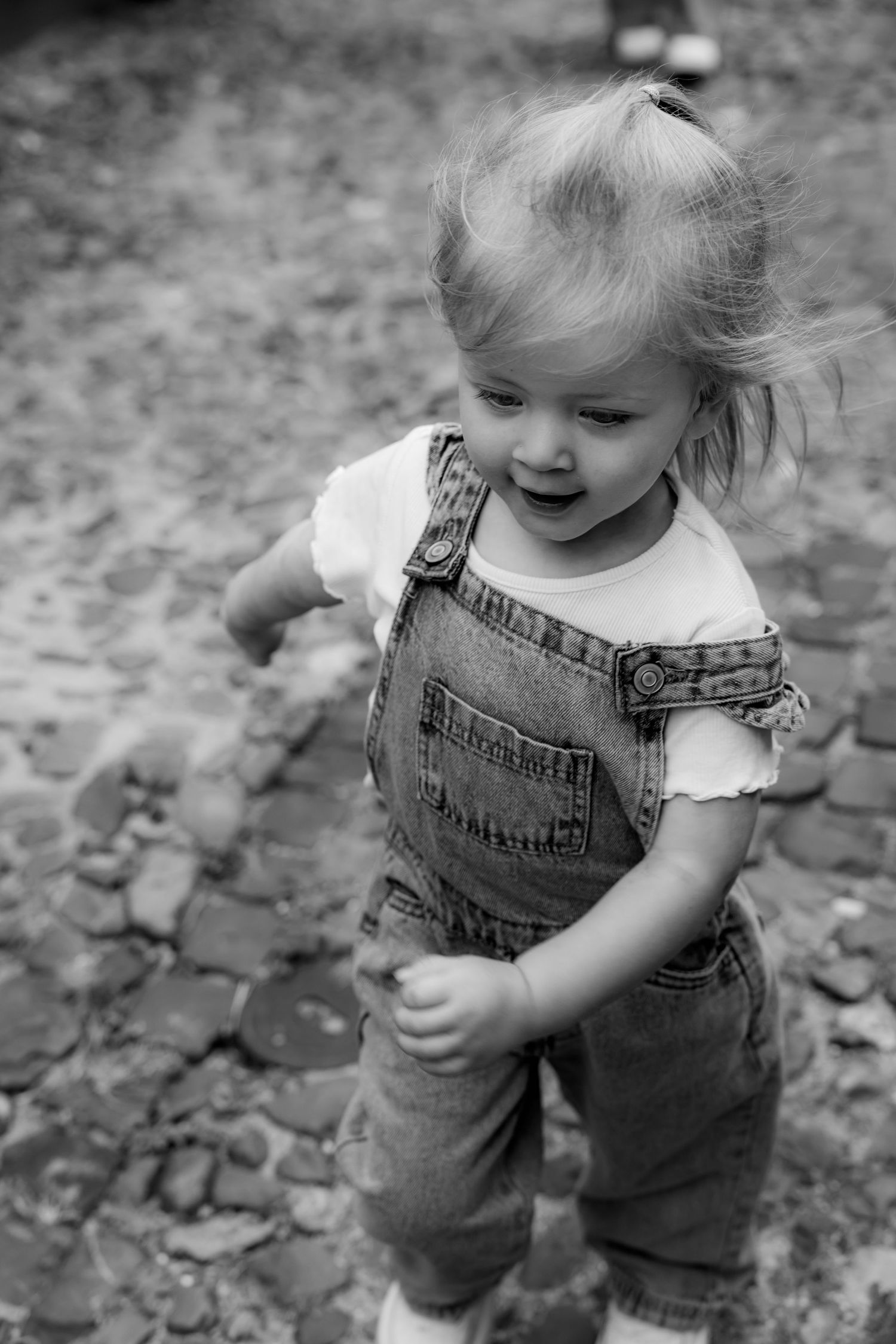 Small child wearing denim overalls plays outdoors in black and white photography.