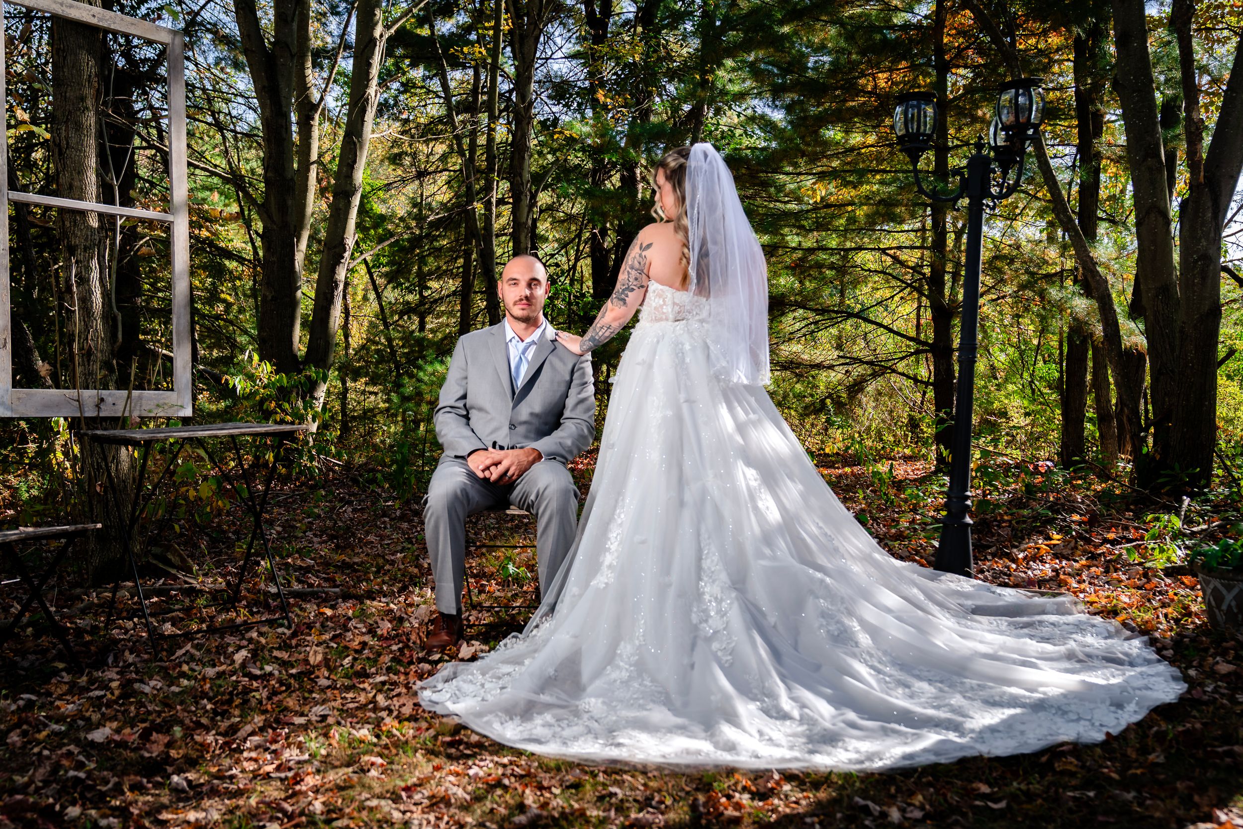 A couple poses for a romantic wedding photo in a wooded setting with autumn leaves on the ground.