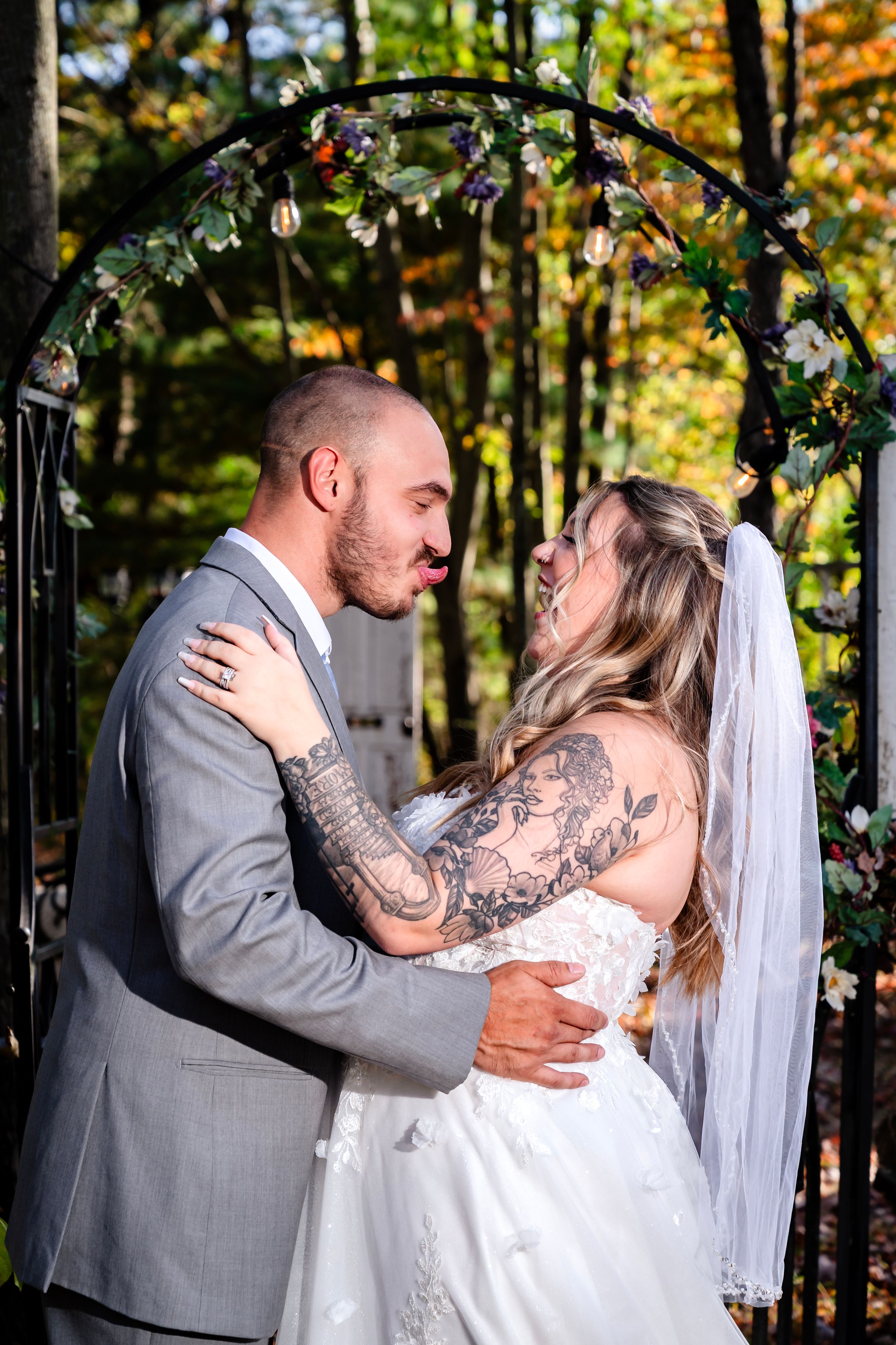 A couple embraces under a floral wedding arch decorated with greenery and lights in an autumn forest.