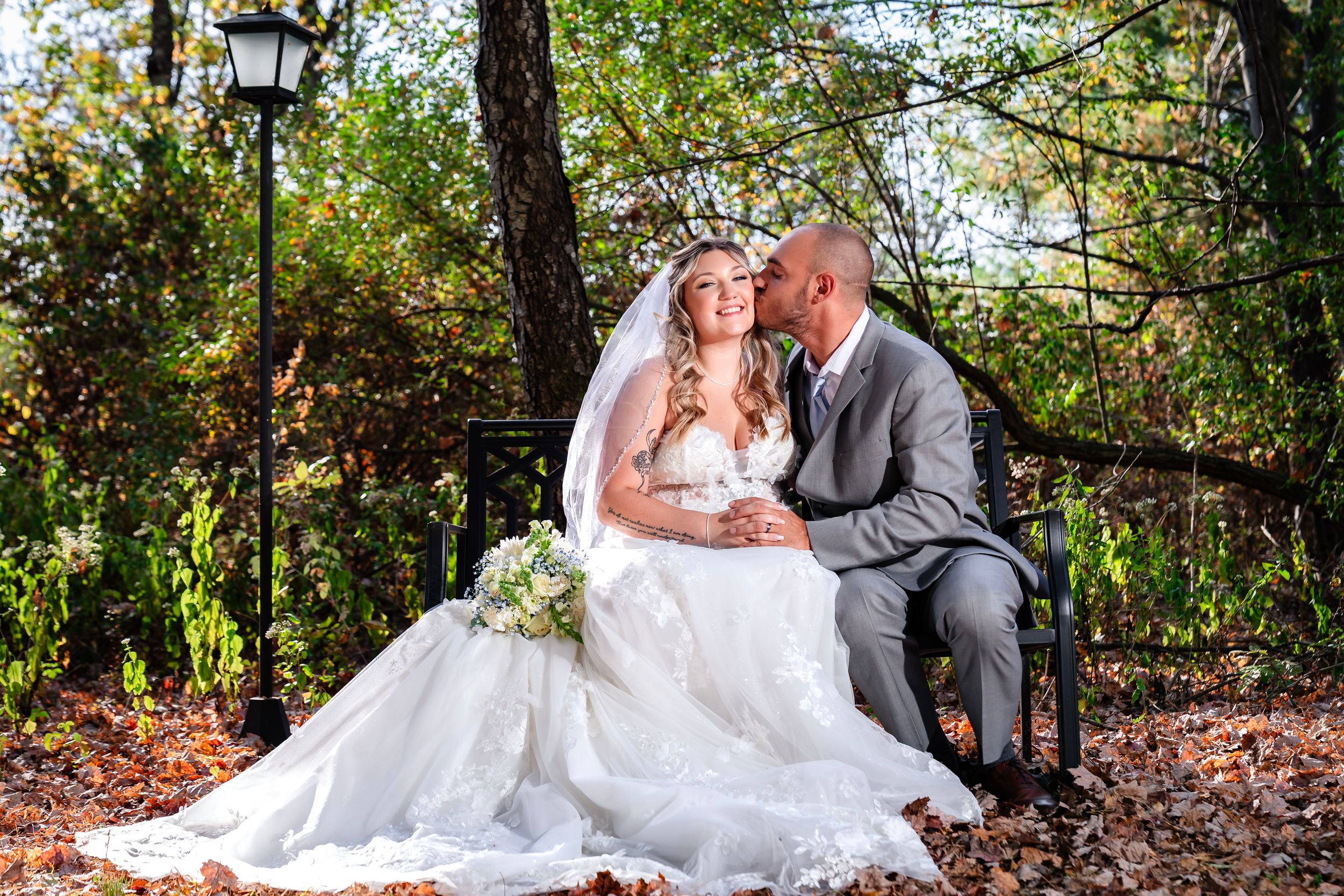 A romantic wedding photo of a couple sitting on autumn leaves in a wooded setting.