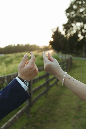 Hands holding up wedding rings against a blurred green outdoor background at sunset.