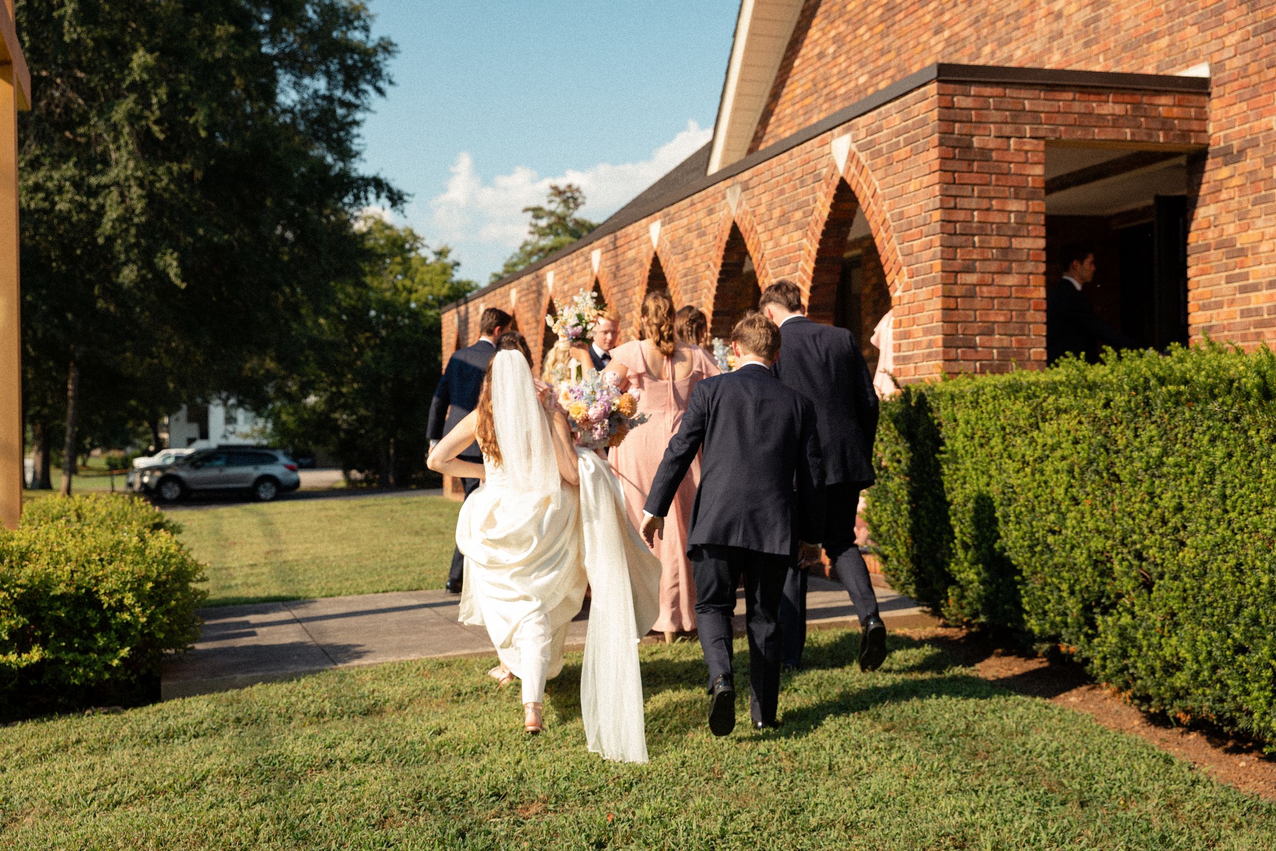 A wedding party walks along a brick building exterior with manicured hedges on a sunny afternoon.