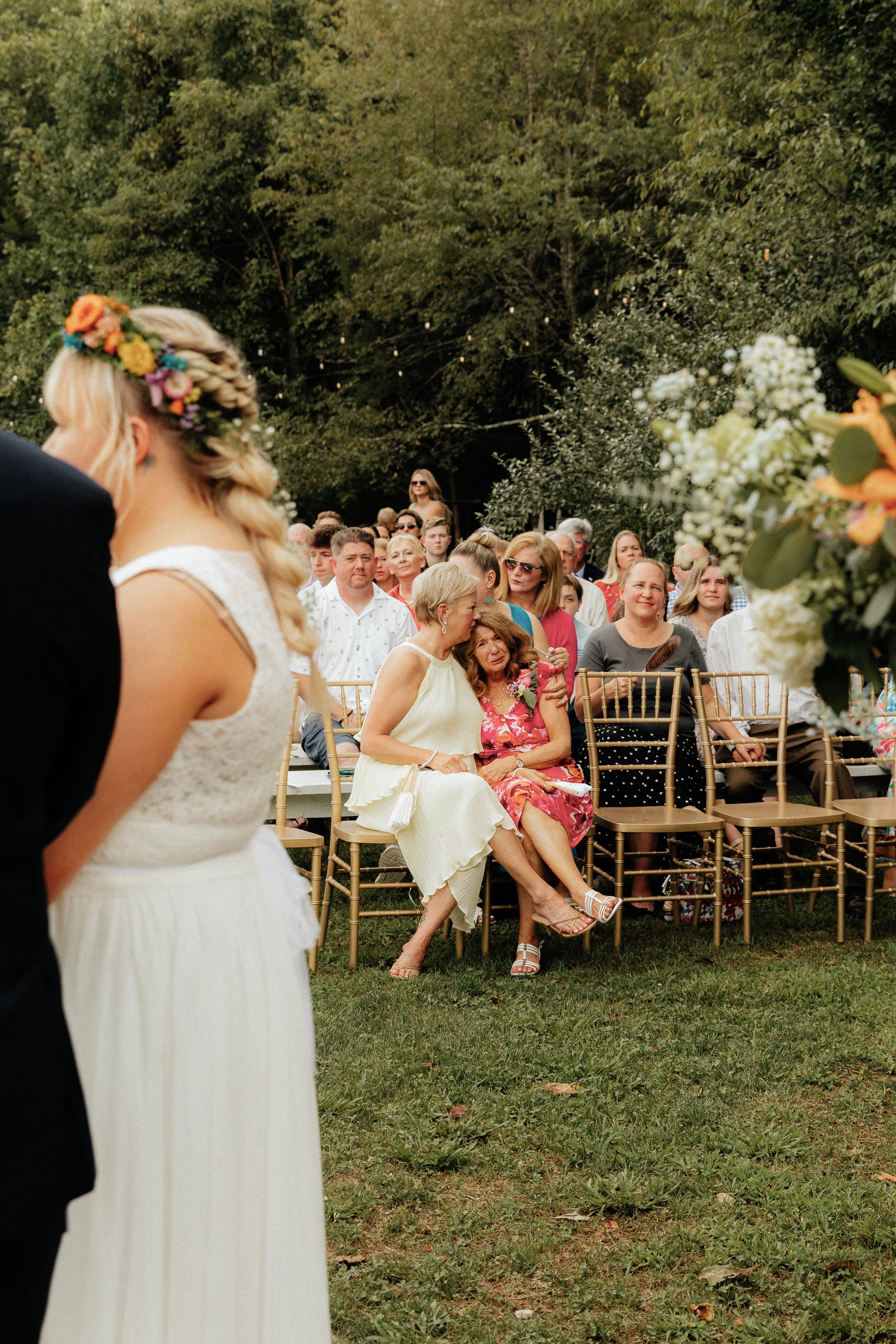 Wedding guests seated on gold chairs laugh together during an outdoor garden ceremony.