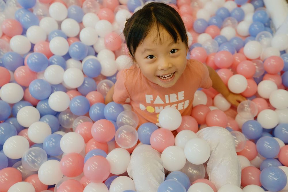 A child plays in a colorful ball pit filled with pink, blue and white plastic balls at an indoor playground.