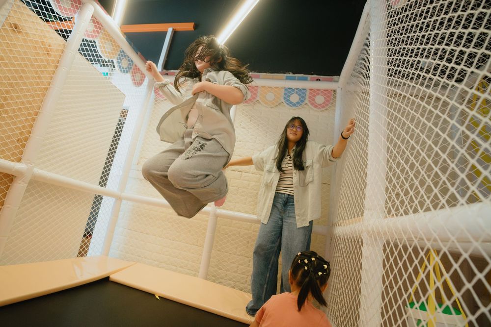 A person practices parkour moves against a white wall with protective netting in an indoor training facility.