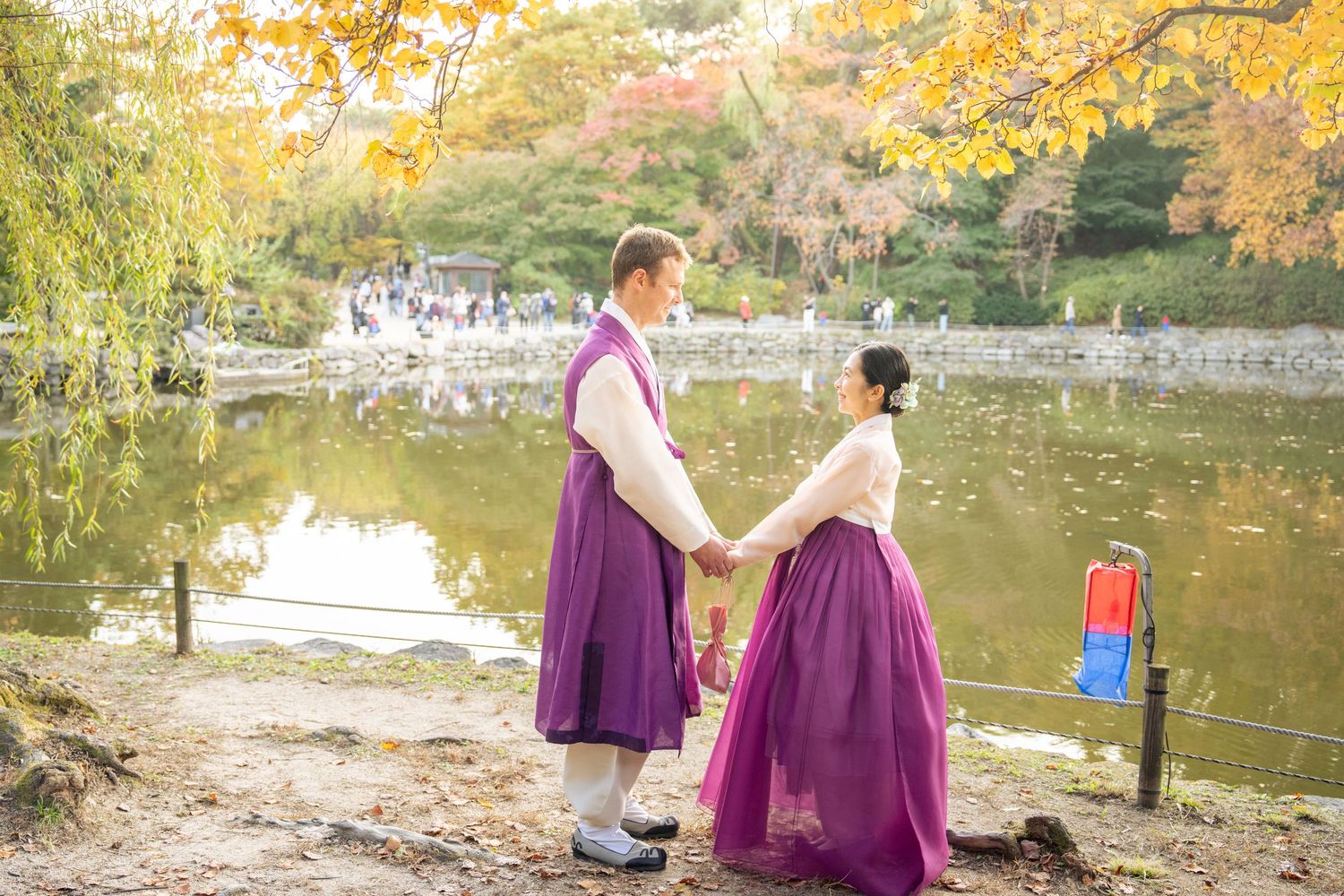 A couple in matching purple outfits poses for photos by a scenic autumn lake during a photoshoot session.