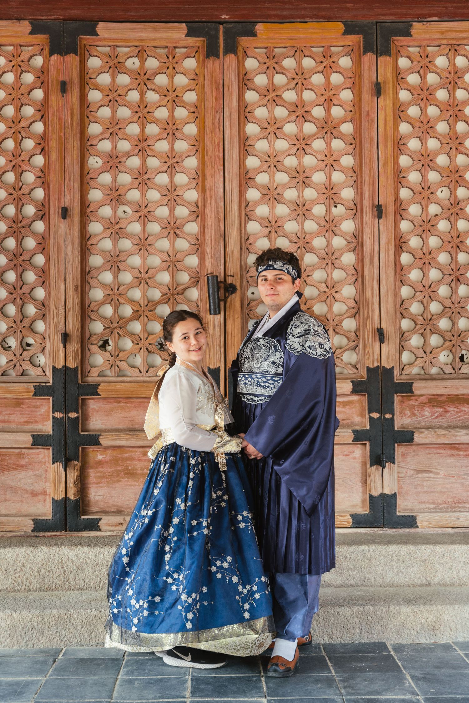 A couple in traditional Korean hanbok dress sit together against an ornate wooden wall with geometric patterns.