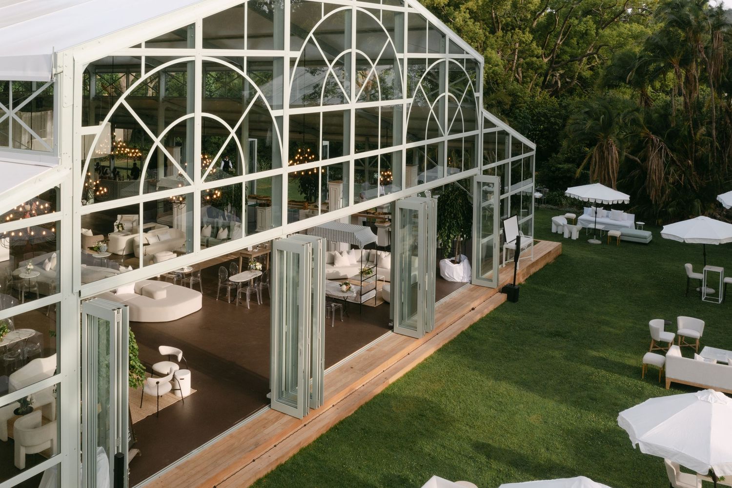 A modern glass conservatory venue with white tables set up for an elegant wedding reception on a manicured lawn.
