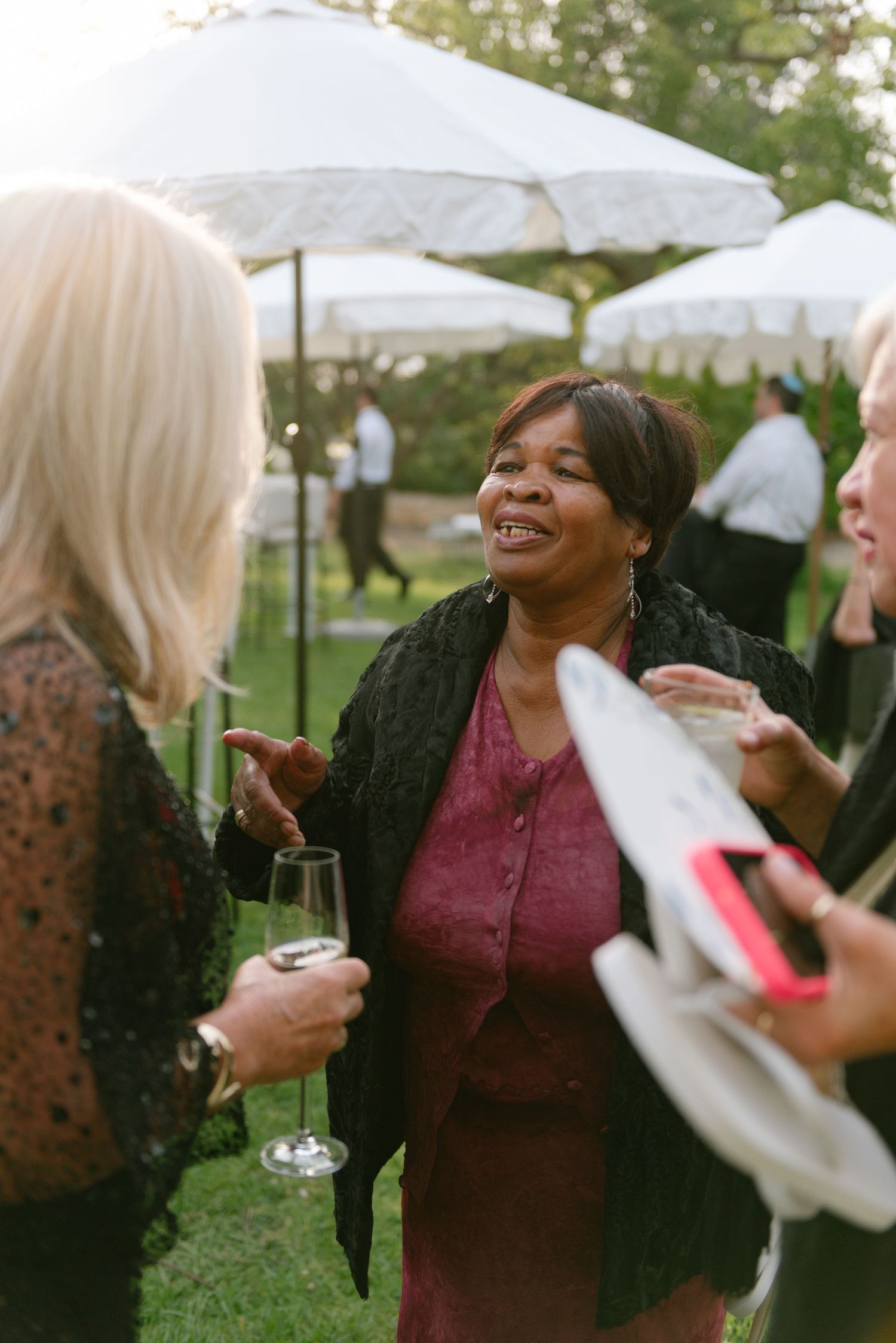 People enjoying conversation and drinks at an outdoor garden party under white umbrellas on a sunny day.