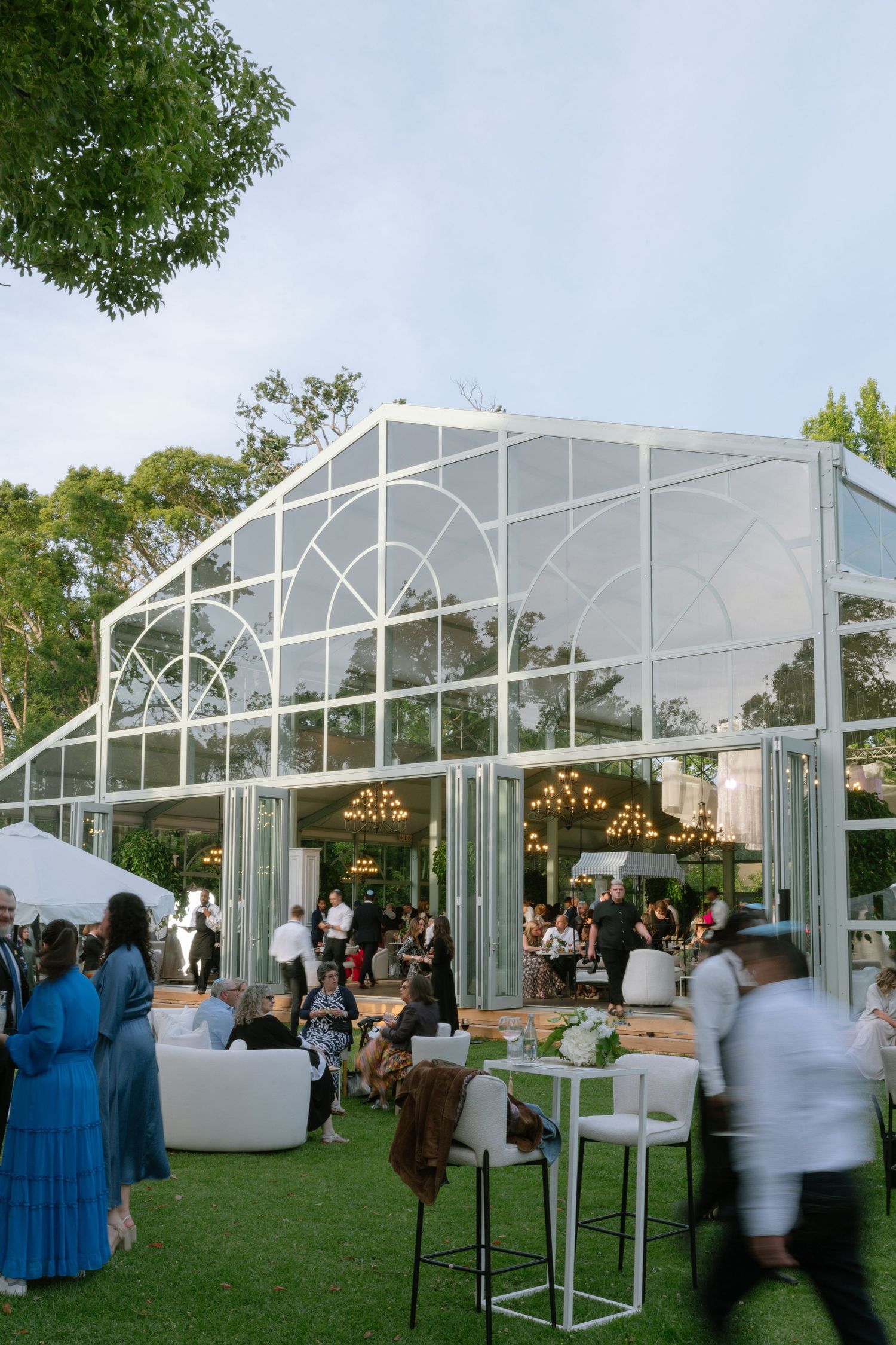 Outdoor wedding reception setup with white tented pavilions and guests mingling on a grassy lawn at dusk.
