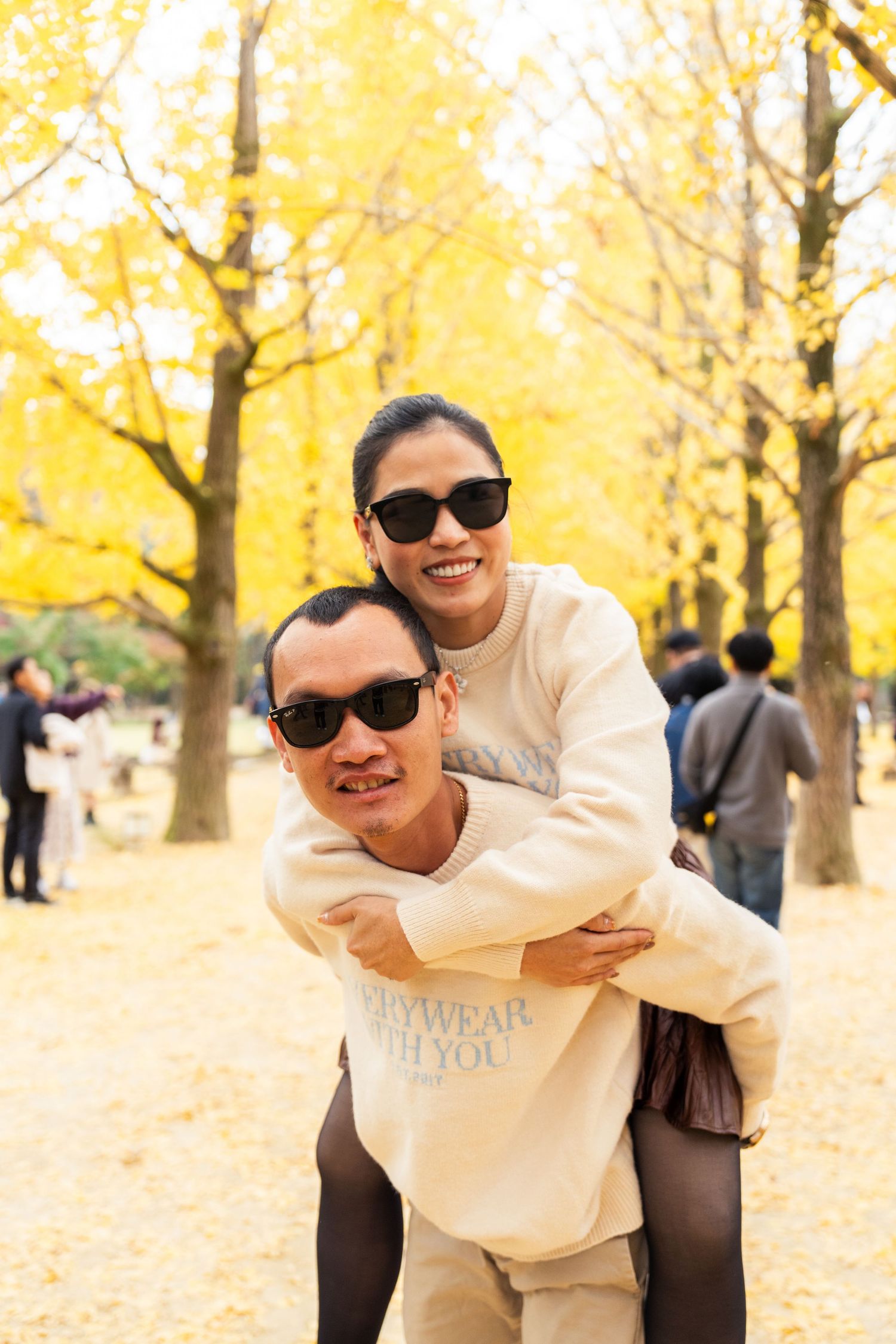 Two people in sunglasses and cream colored outfits share a playful piggyback moment among yellow autumn trees.