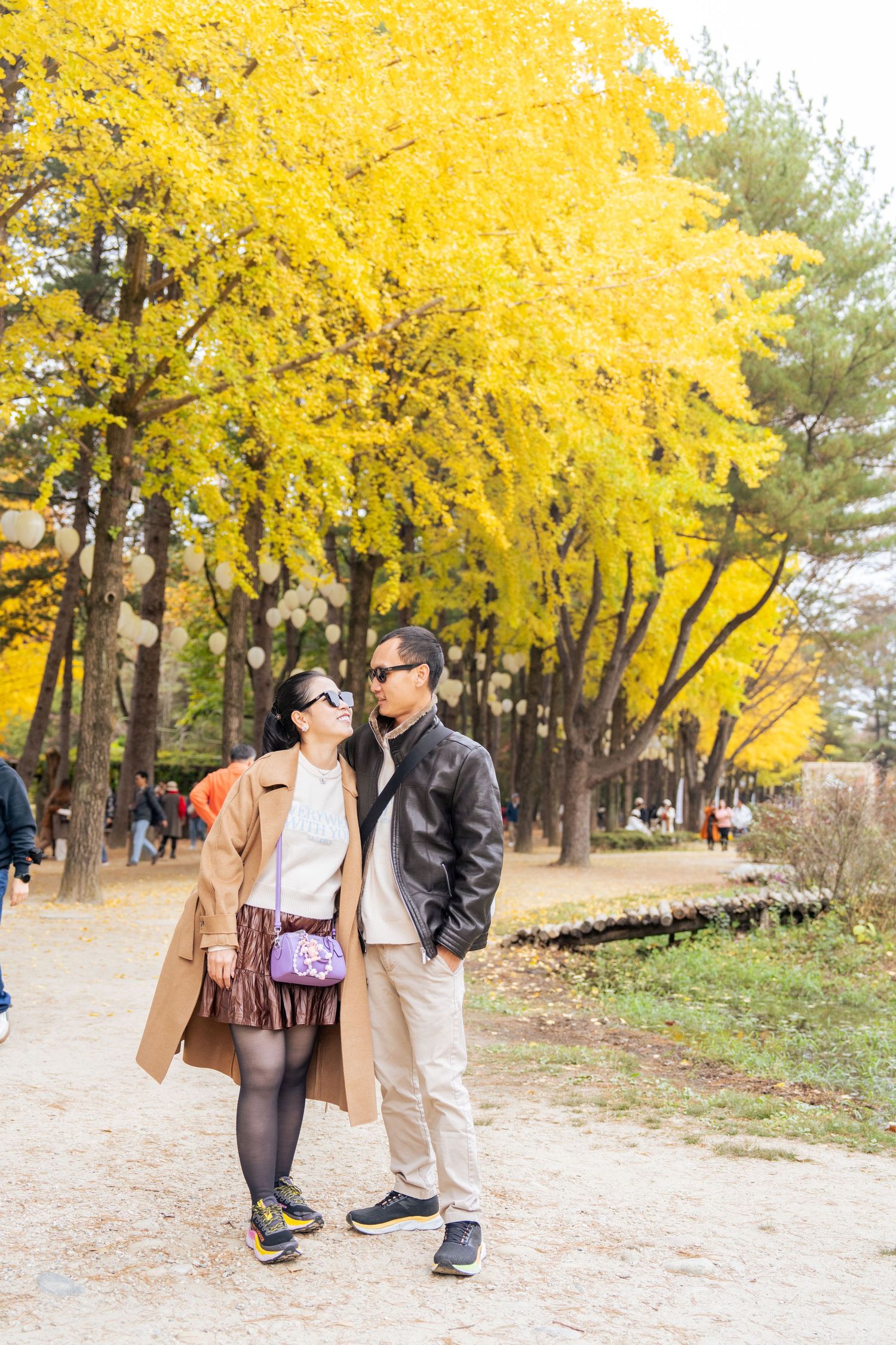A couple shares a romantic moment under vibrant yellow autumn trees along a park pathway.