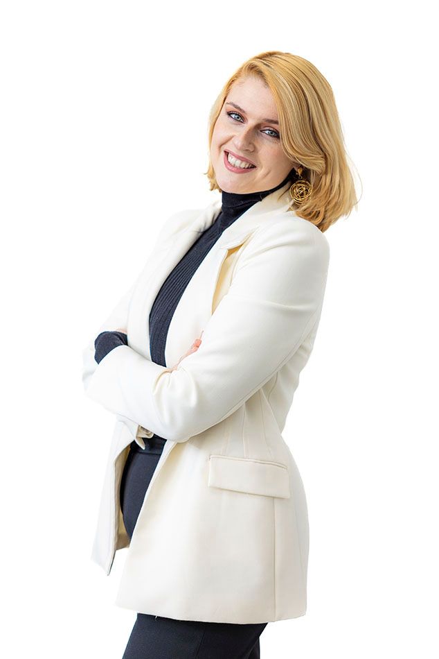 Professional wearing a white blazer and dark top posing confidently against white background.