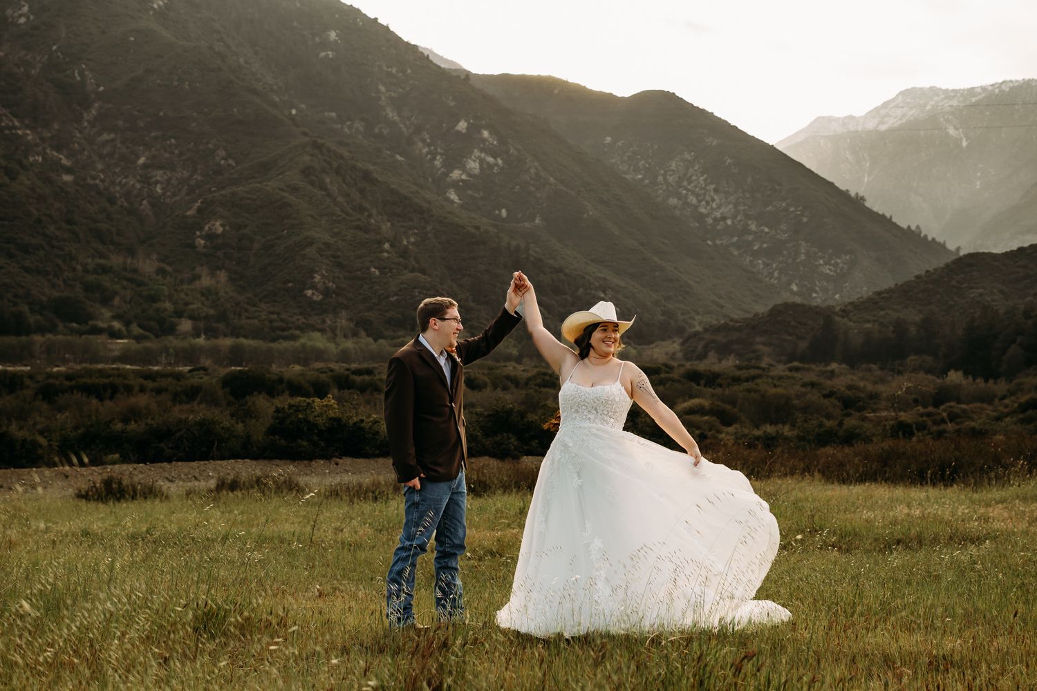 Newlyweds walk along a desert road with mountains in background, bride in white gown and groom in red suit.