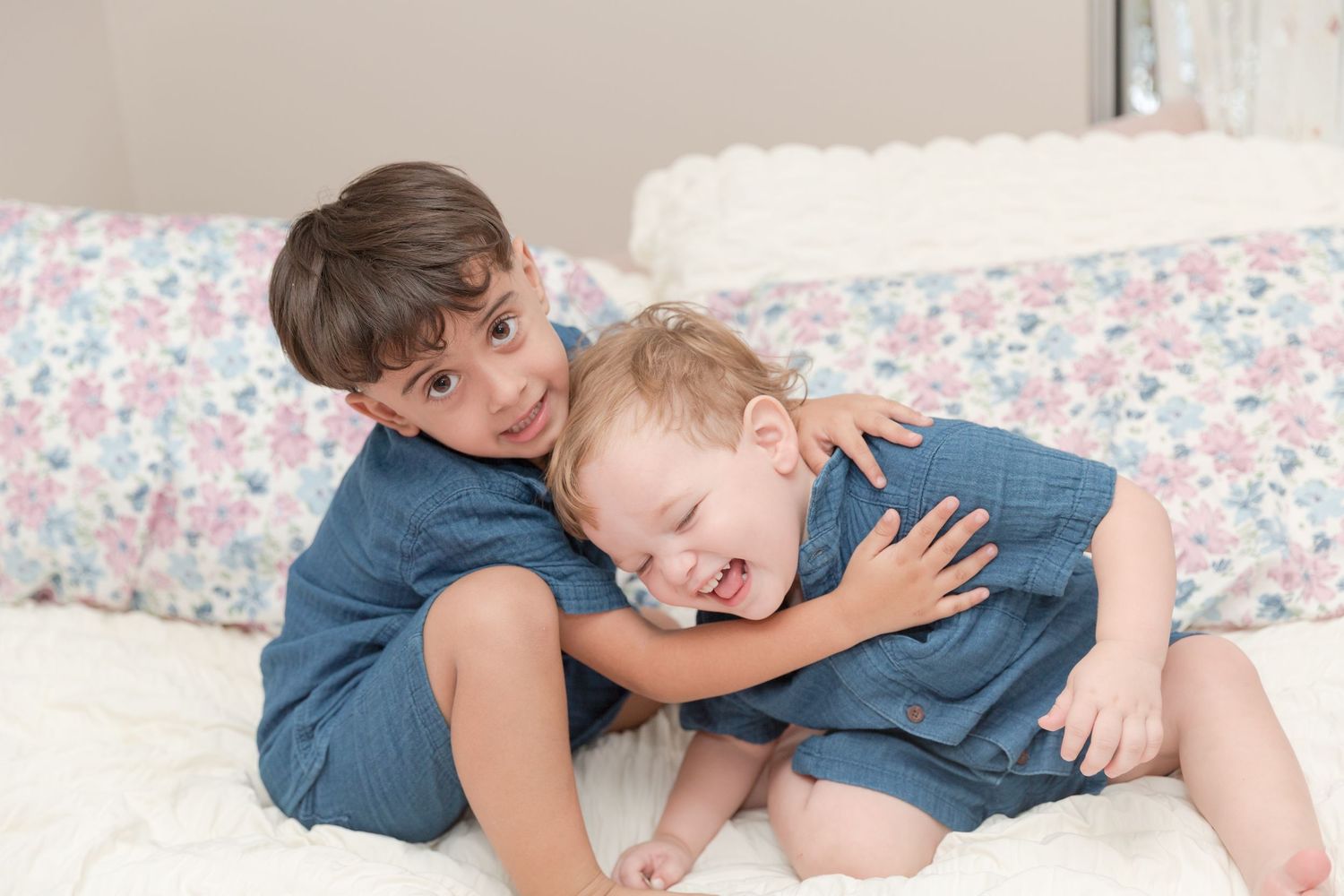 Two young brothers in matching blue outfits laughing and cuddling together on a white bed.