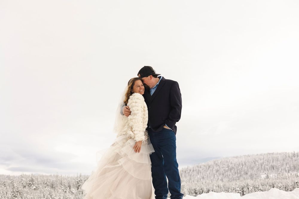 Couple embracing in winter wedding attire with snowy mountain forest backdrop, bride in white gown and groom in dark jacket.