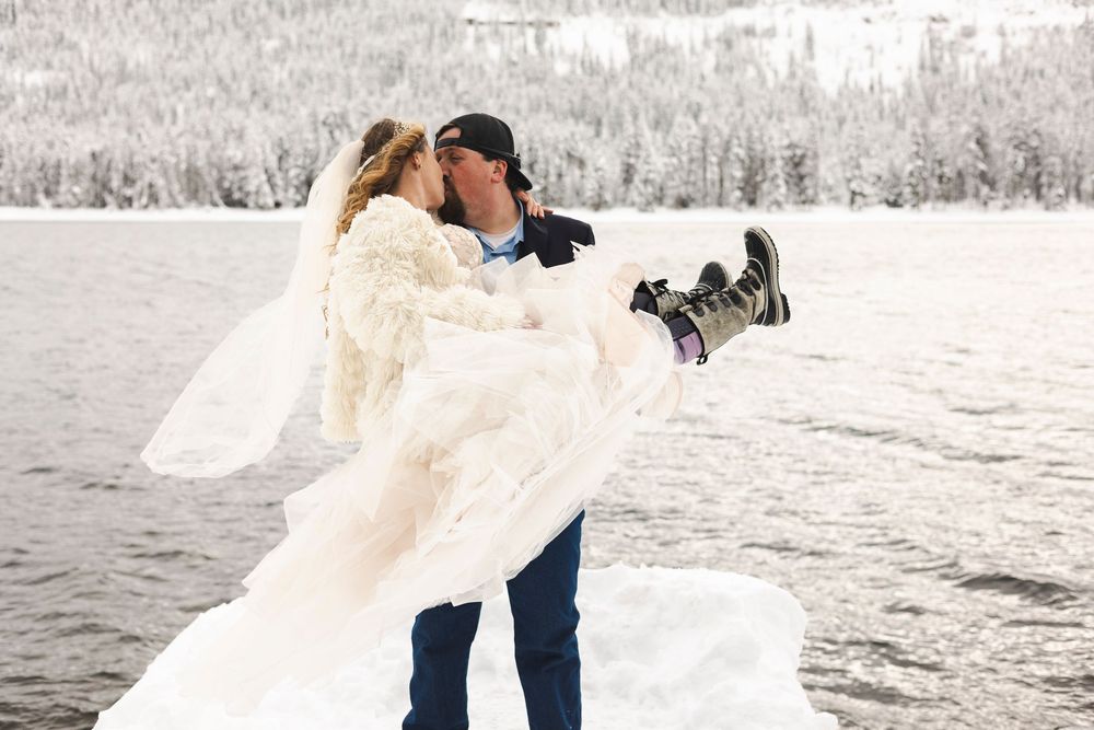 Groom carries bride in wedding dress by snowy lake in winter wonderland wedding photo.