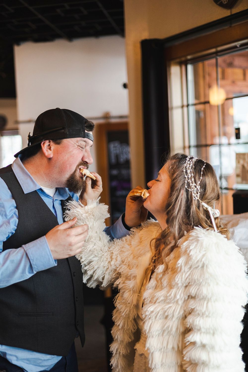 Bride and groom share cake tasting moment indoors, bride wearing feathered jacket and floral headpiece.