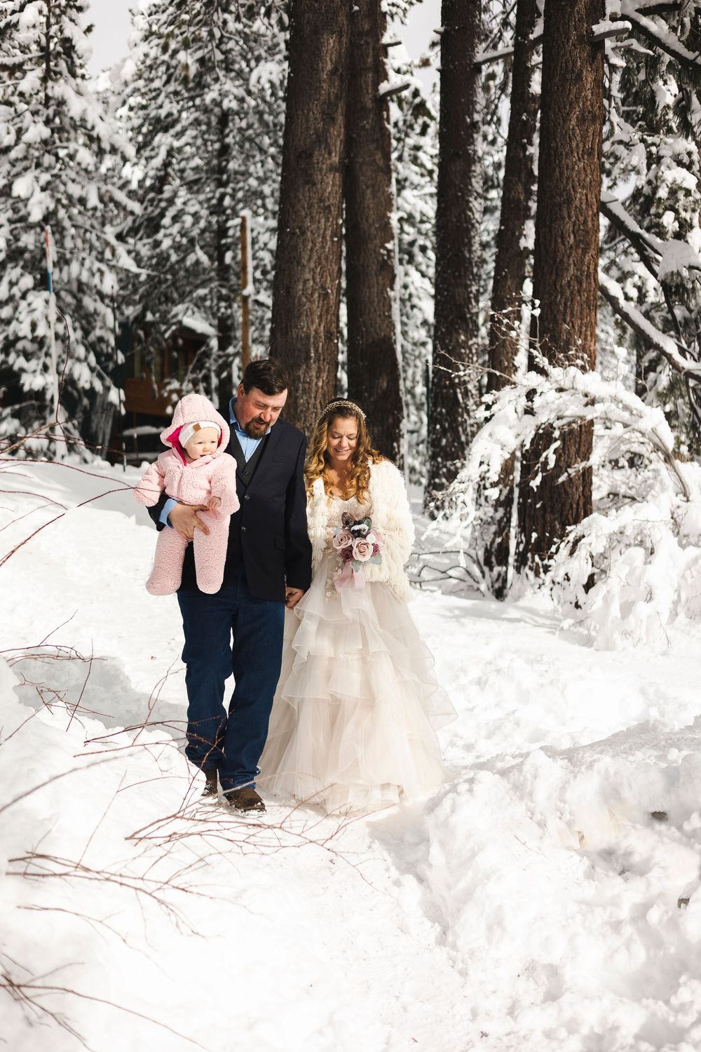 Newlyweds walk through snowy forest with baby after winter wedding ceremony.