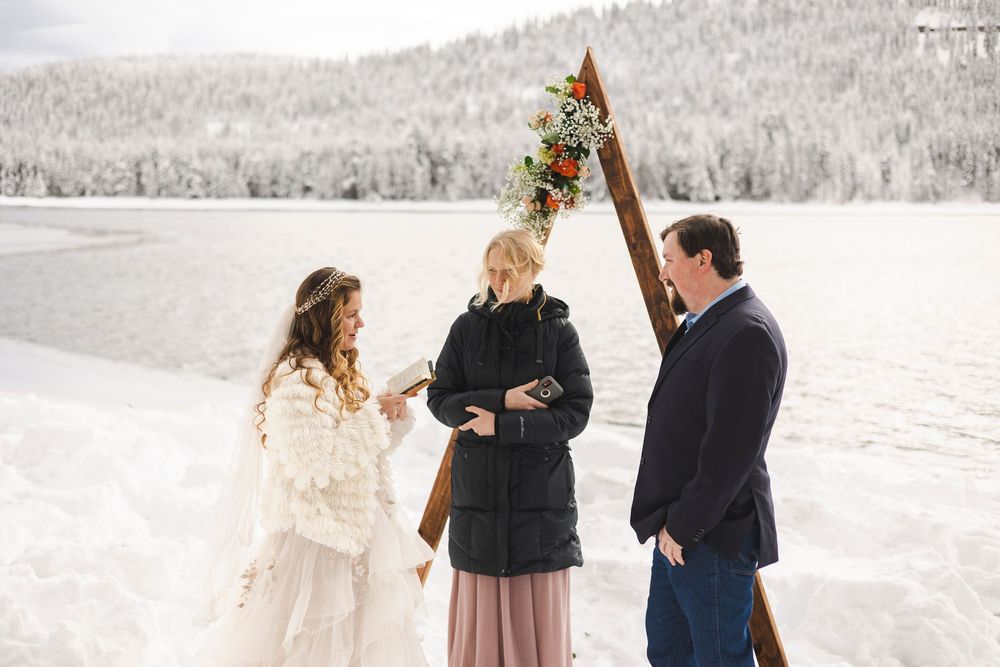 Couple exchanging vows outdoors in snowy winter landscape with officiant at rustic wooden arch wedding altar.