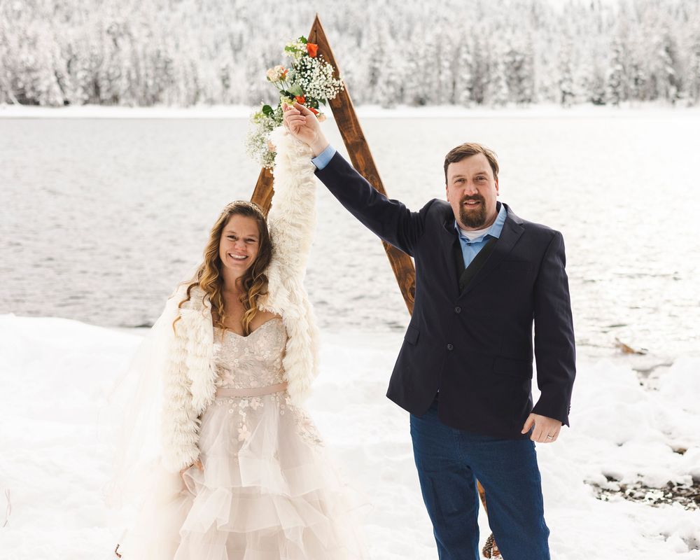 Joyful newlywed couple celebrating winter wedding outdoors in snow, bride in white gown holding bouquet aloft by frozen lake.
