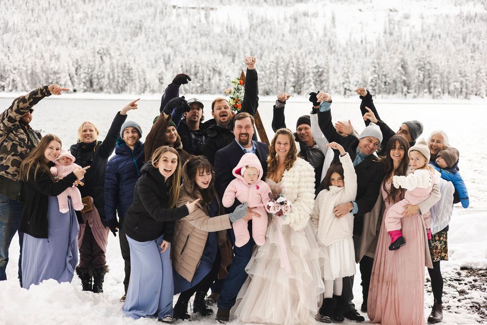 Winter wedding group photo with bride in white gown, groom, and guests celebrating in snowy mountain landscape.