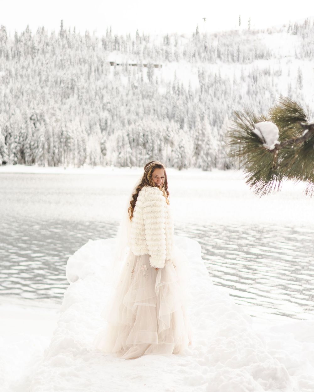 Bride in white tulle skirt and fur jacket standing in snowy winter landscape by frozen lake.