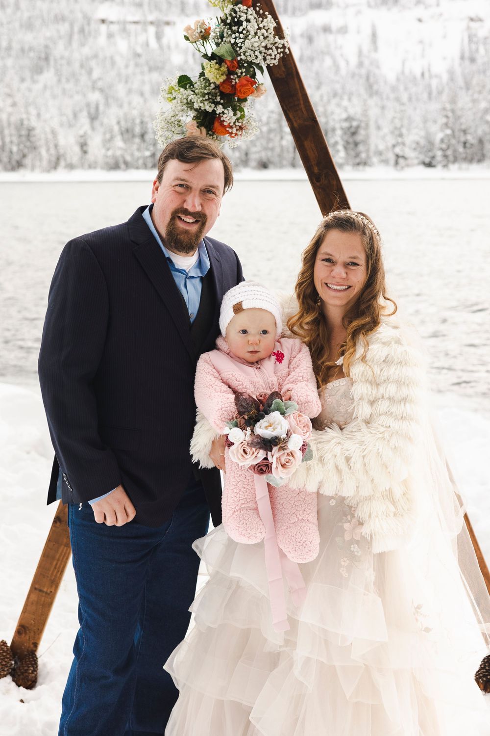 Bride and groom pose with baby at snowy winter wedding ceremony under floral arch.