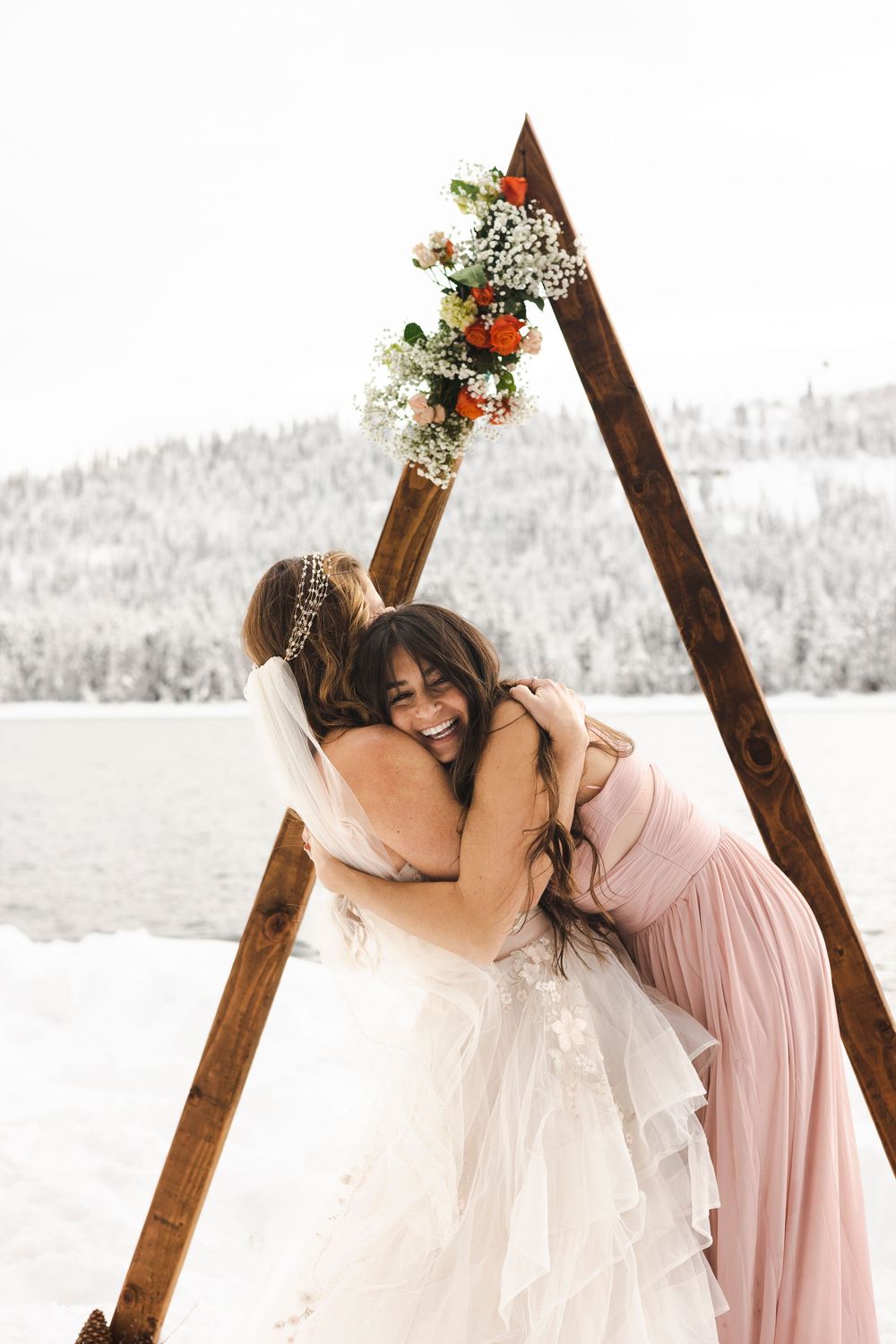 Two brides embracing joyfully under a wooden triangular arch adorned with flowers at a snowy winter wedding.