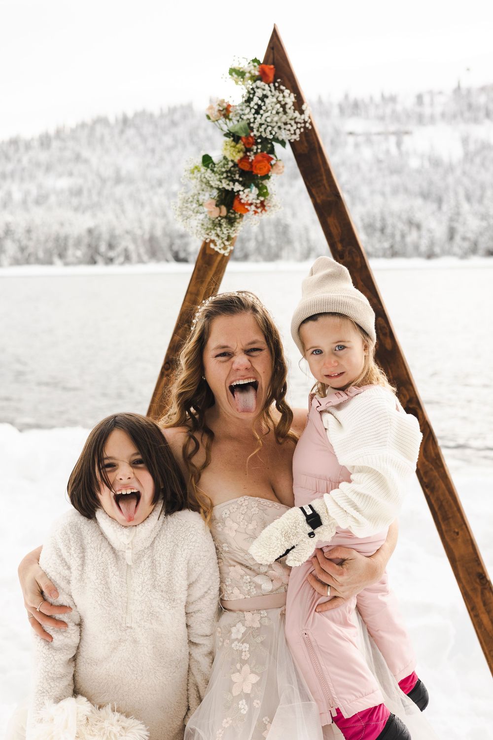 Bride in white gown poses with two young girls, sticking out tongues, under wooden arch with flowers in snowy winter setting.