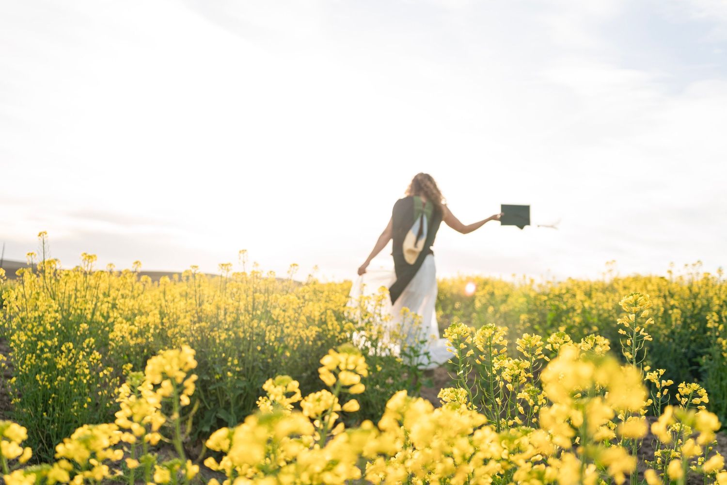 Woman in white dress walking through vibrant yellow flower field, holding a book, with bright sunlight in the background.