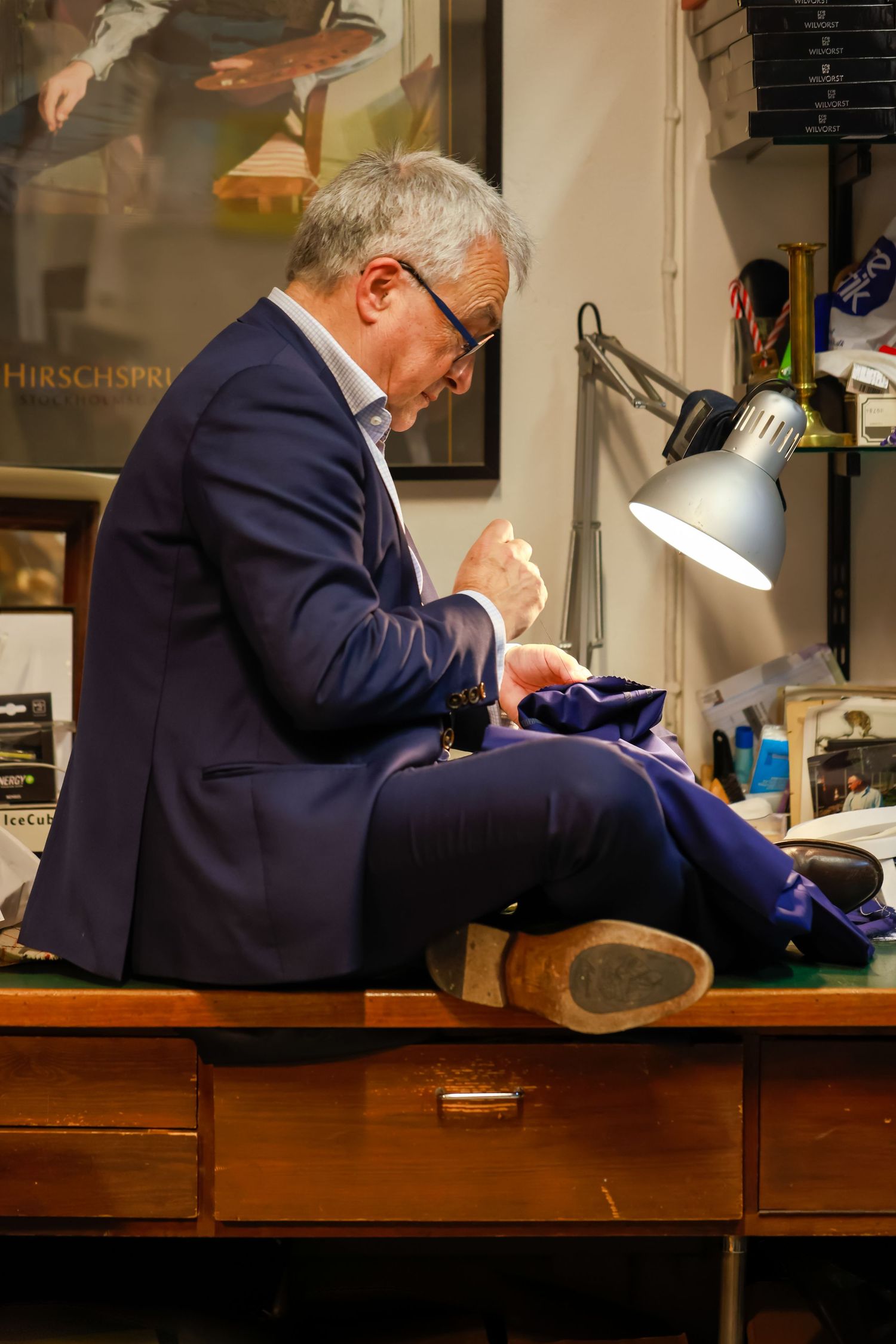 Elderly tailor in blue suit sits on wooden desk, hand-stitching fabric under desk lamp in vintage workshop.