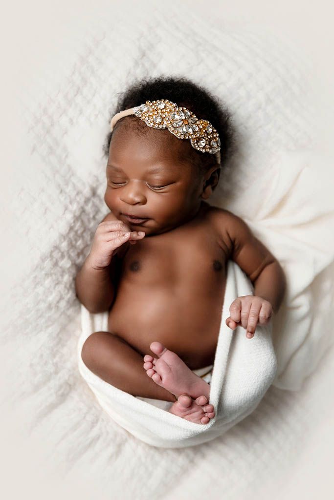 Newborn baby wearing jeweled headband, peacefully sleeping wrapped in white cloth on soft blanket.