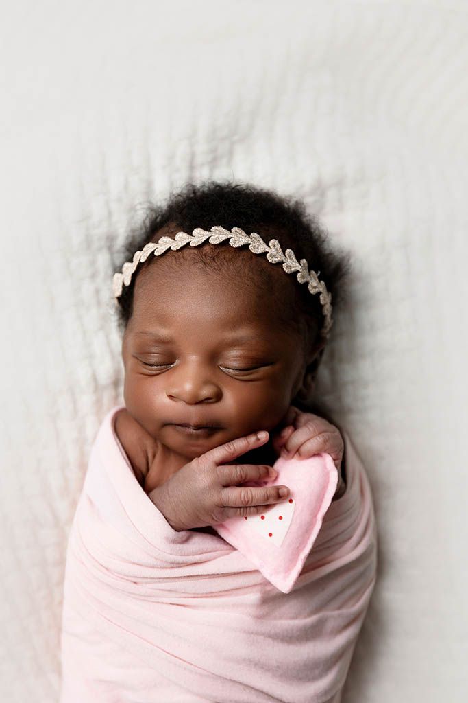 Valentines themed newborn photoshoot of baby girl swaddled in pink wrap with heart headband and a tiny pink heart prop.