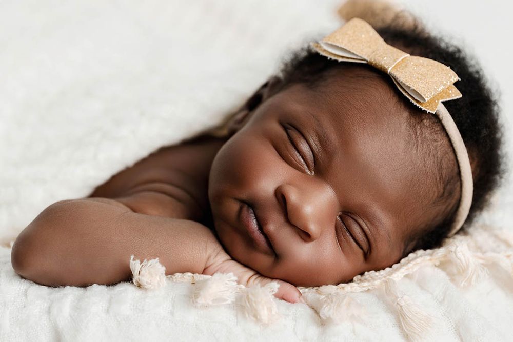 Adorable sleeping newborn baby girl smiling with gold bow headband on soft white blanket.