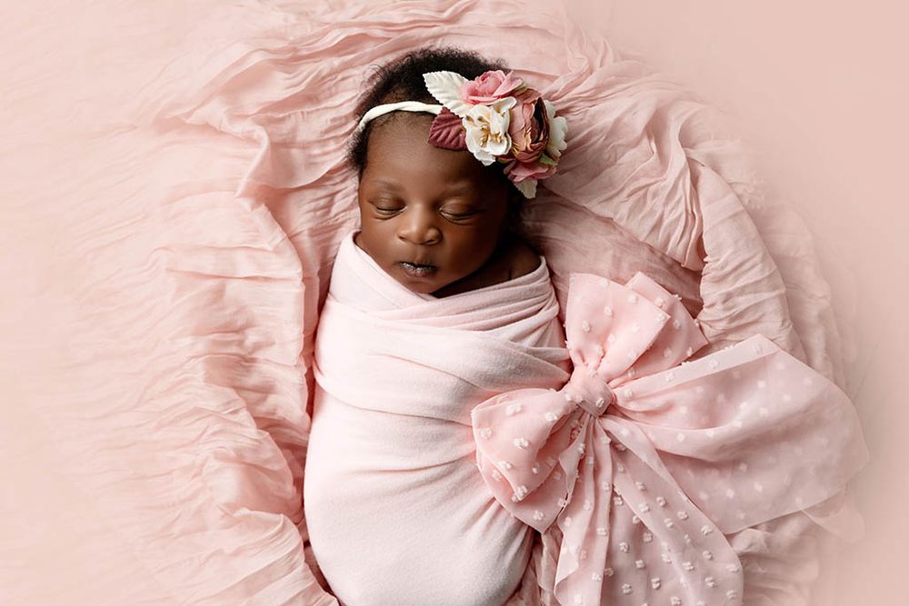 Newborn baby girl swaddled in pink wrap with floral headband sleeping on pink background.