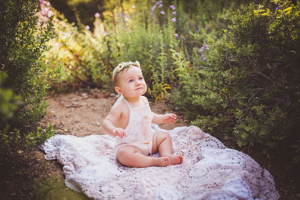 Baby girl in white lace romper sits on blanket in lush garden, looking up with a joyful smile in redlands, ca
