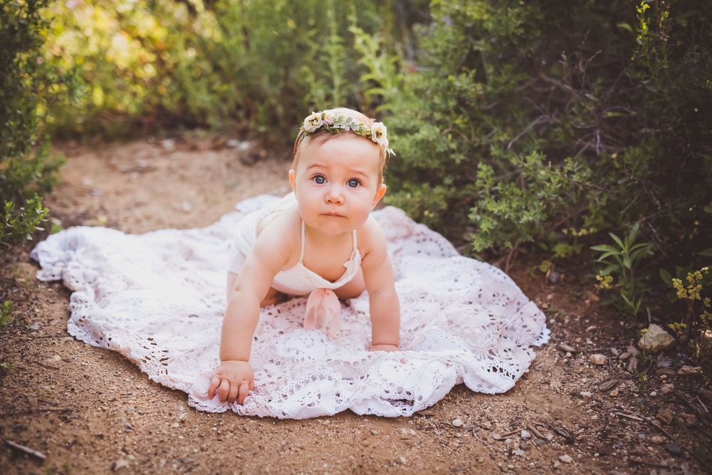 Baby girl in white lace dress and floral headband crawling outdoors on dirt path surrounded by green bushes at a park in redlands, ca